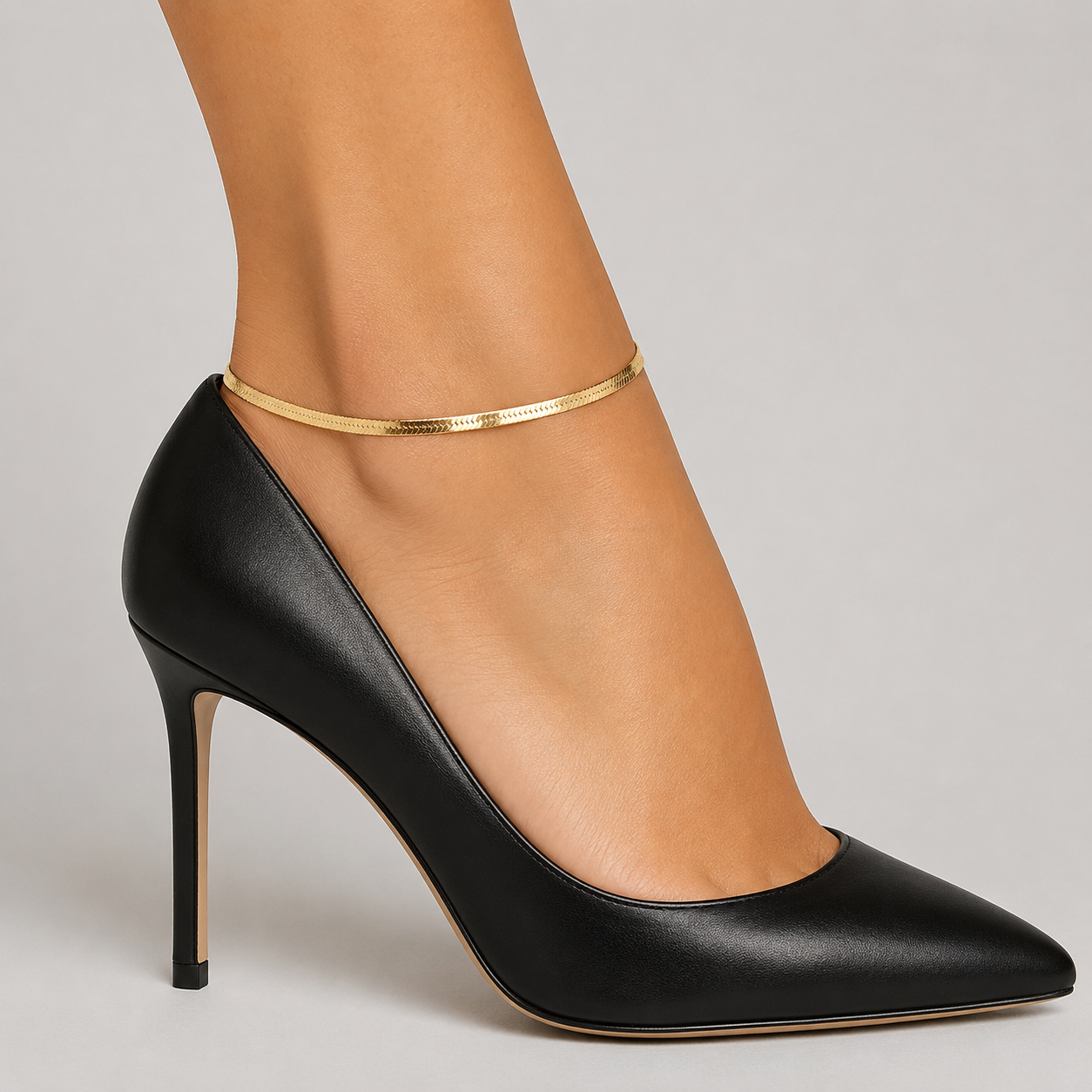 Herringbone flat chain anklet styled with black stiletto heels - sleek gold ankle bracelet, angled view